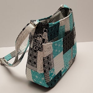 May include: A patchwork shoulder bag in teal, black, white, and grey with a patterned design. It features a long, adjustable strap and two external pockets. The bag is made of fabric.