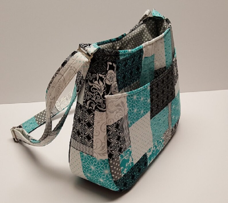 May include: A patchwork shoulder bag in teal, black, white, and grey with a patterned design. It features a long, adjustable strap and two external pockets. The bag is made of fabric.