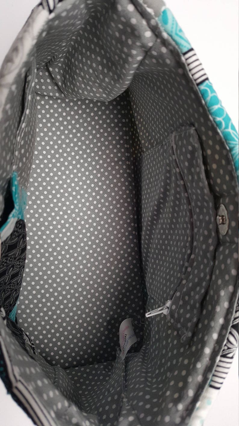 May include: Inside view of a fabric tote bag. The interior lining is grey with white polka dots. The bag has a zipped pocket and a magnetic snap closure. The exterior fabric features patterns in teal, black, and white.
