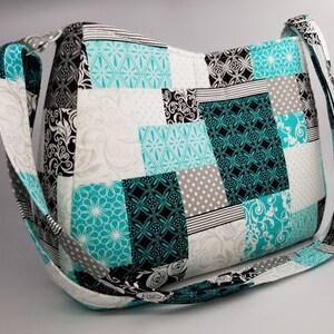 May include: A patchwork shoulder bag in teal, black, white, and grey. The bag has a long strap and is made of quilted fabric. The design incorporates floral, geometric, and polka dot patterns. The bag's measurements are not visible.