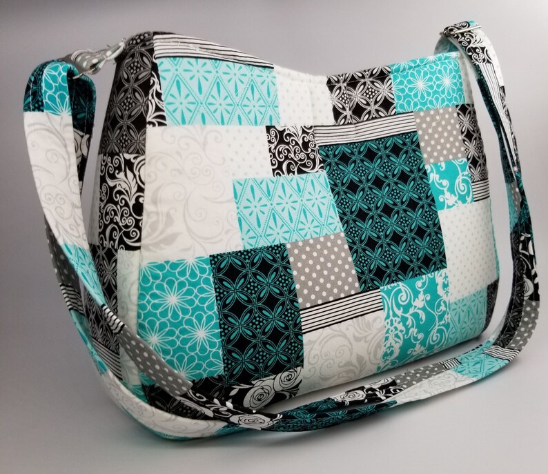 May include: A patchwork shoulder bag in teal, black, white, and grey. The bag has a long strap and is made of quilted fabric. The design incorporates floral, geometric, and polka dot patterns. The bag's measurements are not visible.