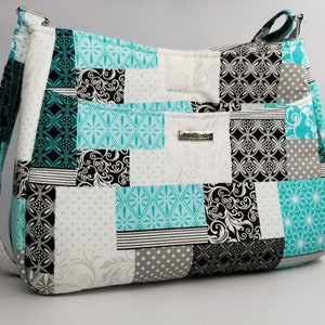 May include: A patchwork shoulder bag featuring squares of patterned fabric in white, black, grey, and teal. The bag has a long strap and a small metal tag that reads "handmade".