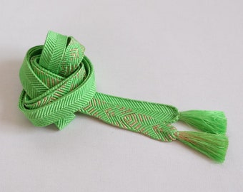 May include: A bright green decorative ribbon with a herringbone pattern and gold accents. The ribbon is tied in a loose knot, with tassels at the ends. The ribbon is likely used for crafts or gift wrapping.