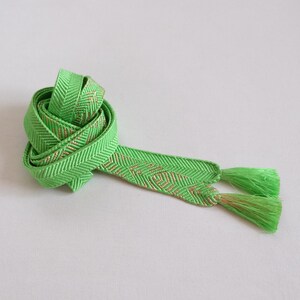 May include: A bright green decorative ribbon with a herringbone pattern and gold accents. The ribbon is tied in a loose knot, with tassels at the ends. The ribbon is likely used for crafts or gift wrapping.
