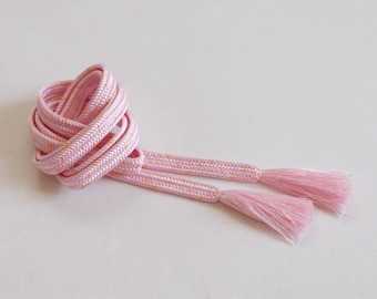 May include: Pink and orange cord with tassels. The cord is made of a thin, braided material and has a subtle pattern of small orange dots. The tassels are made of the same material as the cord and are about 2 inches long.