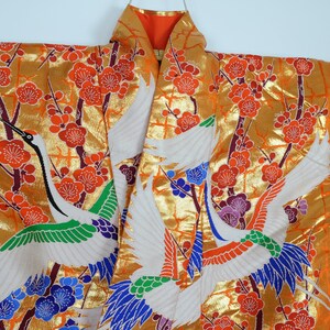 May include: A traditional Japanese kimono with a gold background and a pattern of white cranes flying among red and purple flowers. The kimono is made of silk and has a luxurious feel.