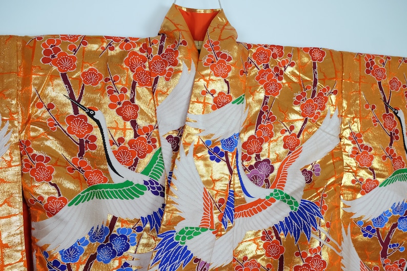 May include: A traditional Japanese kimono with a gold background and a pattern of white cranes flying among red and purple flowers. The kimono is made of silk and has a luxurious feel.