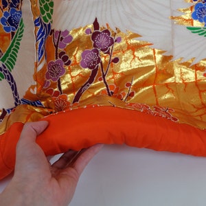 May include: A traditional Japanese kimono with a white background and gold, red, green, and blue floral and crane patterns. The kimono has a red lining and is folded in half.