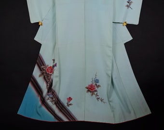 May include: A light blue kimono with a floral pattern of pink and red roses, blue flowers, and brown branches. The kimono has a blue and brown ombre design on the bottom hem.