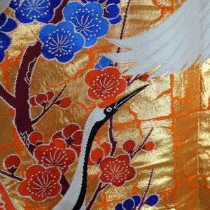 May include: A white fabric with a gold background and a pattern of red and blue flowers and white cranes. The fabric has a textured, woven look.