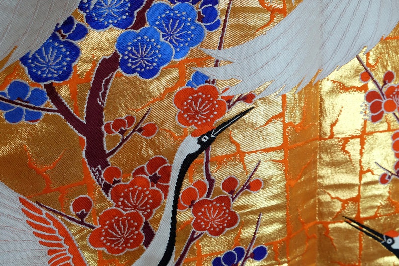 May include: A white fabric with a gold background and a pattern of red and blue flowers and white cranes. The fabric has a textured, woven look.