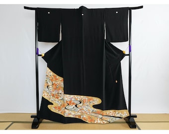 Op de afbeelding: Een zwarte kimono met een gouden en oranje bloemenpatroon aan de zoom. De kimono hangt aan een zwarte standaard.