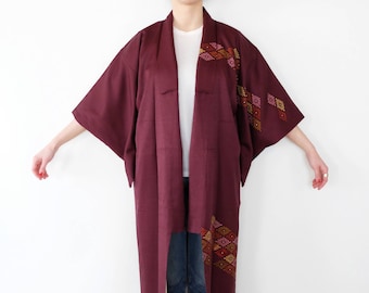 Op de afbeelding: Een bordeauxrode kimono met een geometrisch patroon in tinten roze, oranje en goud. De kimono is open aan de voorkant, waardoor een wit onderhemd zichtbaar is.