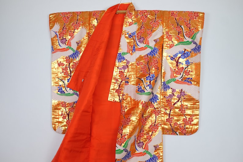 May include: A traditional Japanese kimono with a gold background and a pattern of white cranes flying among pink cherry blossoms. The kimono is lined with a bright orange fabric.