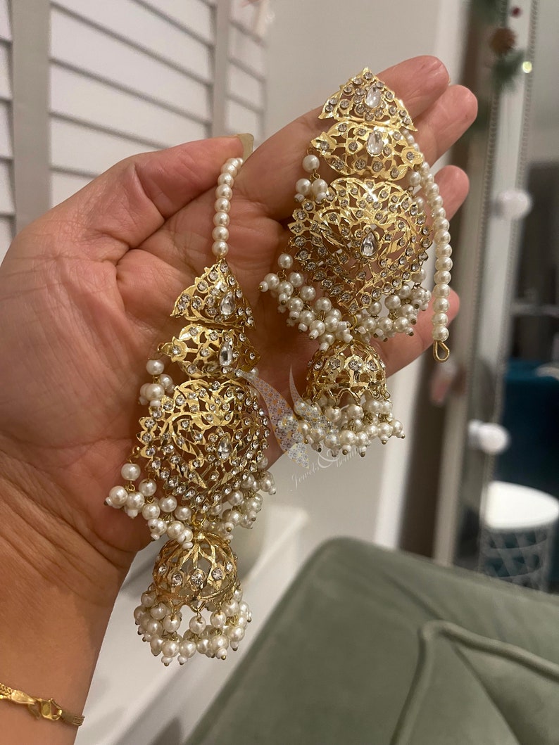 May include: Gold earrings with intricate designs and white pearl accents. The earrings feature a dangling design with multiple layers of pearls.