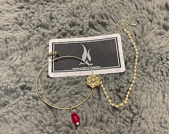 May include: Gold nose ring with a flower design and a dangling red stone. The ring is attached to a chain with small white beads.
