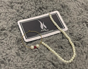 May include: A gold hoop earring with three white pearls and a red gemstone. A strand of white pearls is also pictured on a white card with the text "Jewels & Beauty - A blend of trend and tradition."
