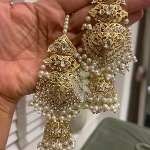May include: A pair of gold earrings with intricate designs and white pearl accents. The earrings feature a dangling bell-shaped charm with a floral pattern.