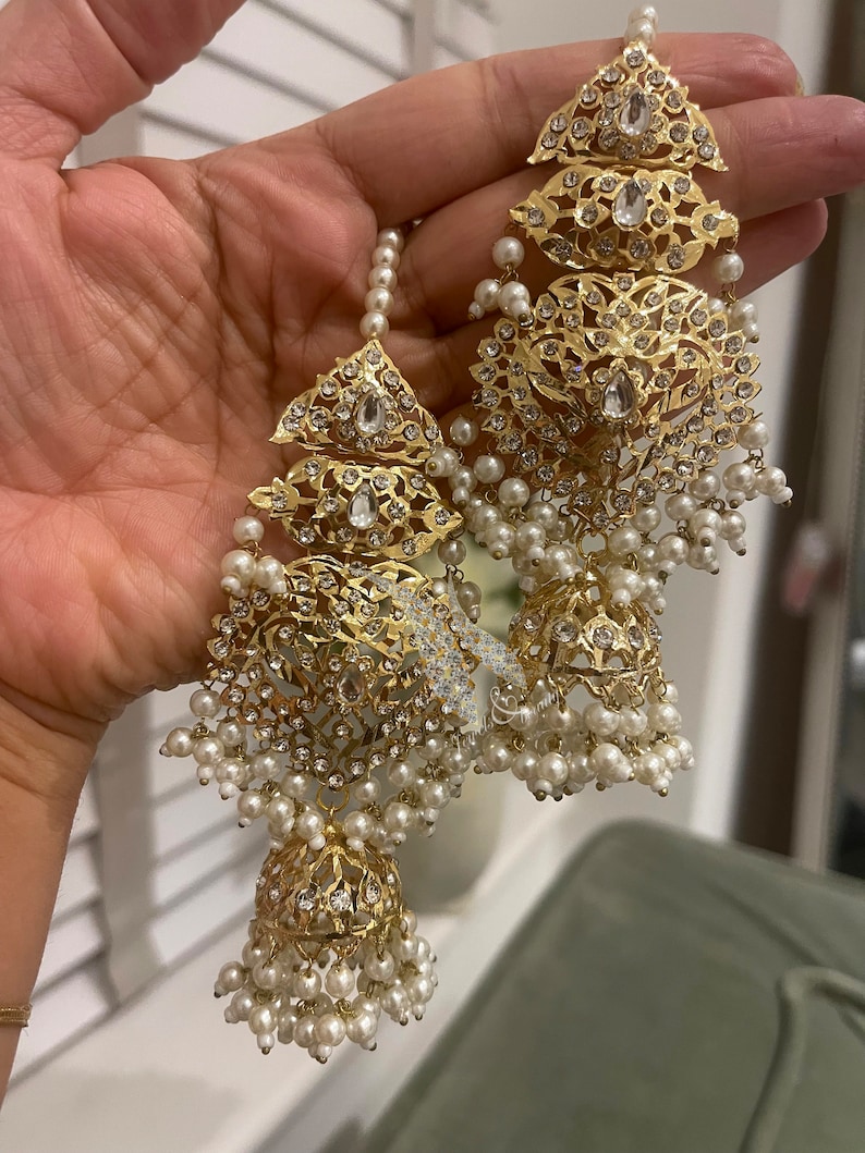 May include: A pair of gold earrings with intricate designs and white pearl accents. The earrings feature a dangling bell-shaped charm with a floral pattern.