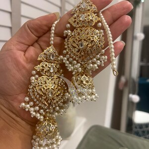 May include: A pair of gold earrings with intricate designs and white pearl accents. The earrings feature a large, round, ornate pendant with a smaller, round pendant hanging below it. The earrings are adorned with small, white pearls.