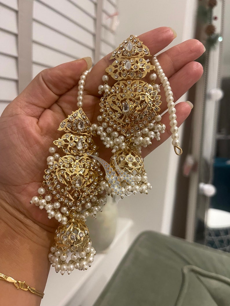 May include: A pair of gold earrings with intricate designs and white pearl accents. The earrings feature a large, round, ornate pendant with a smaller, round pendant hanging below it. The earrings are adorned with small, white pearls.