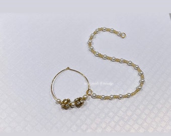 May include: Gold nose ring with a pearl chain. The nose ring has a small, round, jeweled design.