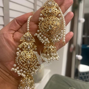 May include: A pair of gold earrings with intricate designs and white pearl accents. The earrings feature a dangling bell-shaped charm with a pearl drop.