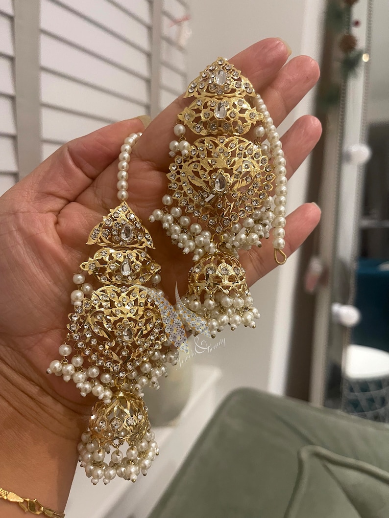 May include: A pair of gold earrings with intricate designs and white pearl accents. The earrings feature a dangling bell-shaped charm with a pearl drop.