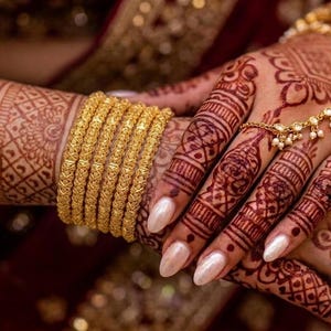 May include: A set of five textured gold bangles worn on the wrist, accompanied by a delicate gold chain bracelet featuring small dangling beads. The skin is decorated with intricate reddish-brown henna patterns, and the fingernails are painted a pearlescent white.