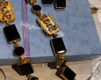 May include: A gold necklace with black rectangular and round stones. The necklace is on a blue background with a purple ribbon. The necklace is paired with matching earrings. The text "tulip jewels & beauty" is visible on the background.