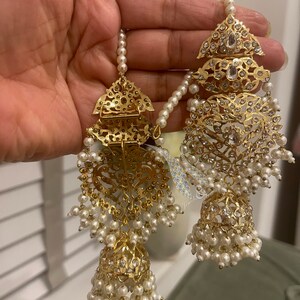 May include: A pair of gold earrings with intricate designs and white pearl accents. The earrings feature a large, ornate pendant with a smaller pendant hanging below it. The earrings are adorned with small, sparkling stones.