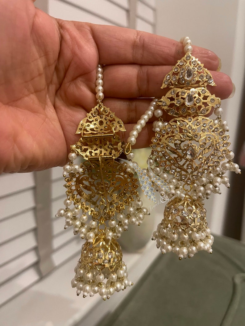 May include: A pair of gold earrings with intricate designs and white pearl accents. The earrings feature a large, ornate pendant with a smaller pendant hanging below it. The earrings are adorned with small, sparkling stones.