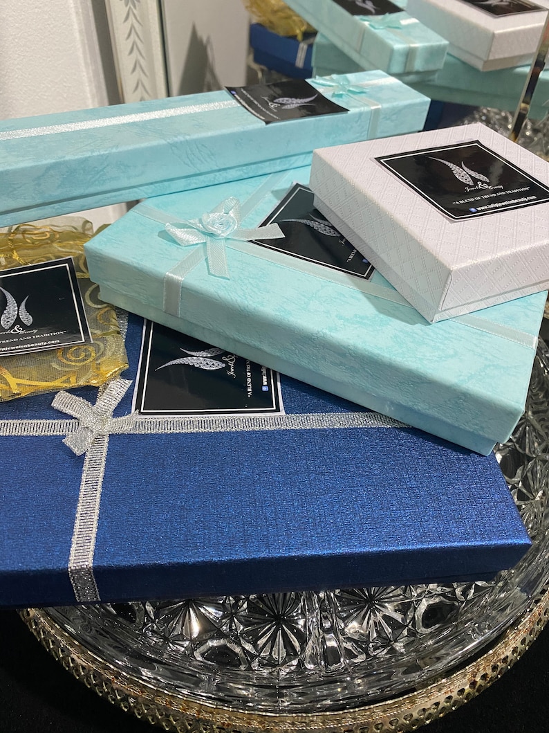 May include: Three blue, white, and light blue gift boxes with a black and white logo that says "Jewelry Beauty - A Blend of Tradition & Modernity" and a website address. The boxes are stacked on top of each other and are sitting on a clear glass plate.