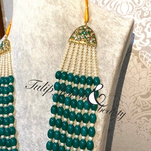 May include: An ornate necklace with multiple strands of green beads and white pearls. The necklace features gold-colored accents with green and clear gemstones. The text "Tulip Jewels & Beauty" is visible on the image.