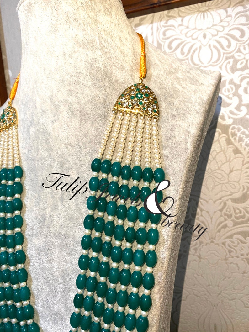 May include: An ornate necklace with multiple strands of green beads and white pearls. The necklace features gold-colored accents with green and clear gemstones. The text "Tulip Jewels & Beauty" is visible on the image.