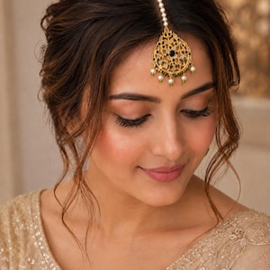 May include: A gold-toned maang tikka headpiece featuring a teardrop-shaped pendant with intricate filigree, embedded with small green stones and clear crystals, accented by dangling white pearls and a single central black stone on a pearl chain.