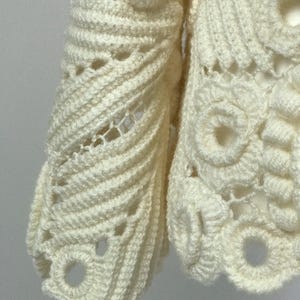 May include: Close-up of a cream-colored, open-weave crochet garment. The intricate design features textured patterns, including spiral details and circular motifs. The handmade piece showcases a delicate, airy construction.