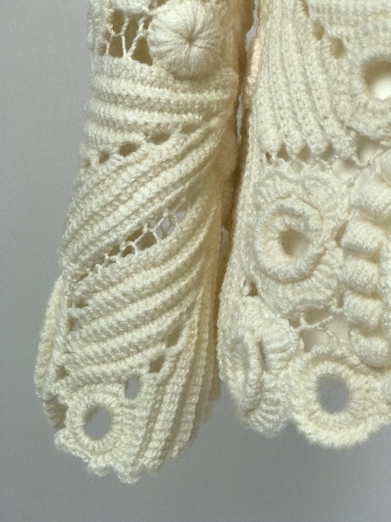 May include: Close-up of a cream-colored, open-weave crochet garment. The intricate design features textured patterns, including spiral details and circular motifs. The handmade piece showcases a delicate, airy construction.