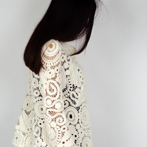 May include: Ivory-colored crochet top with intricate circular and floral patterns. The openwork design features a high collar and long sleeves. The top is made of a soft, lightweight material.