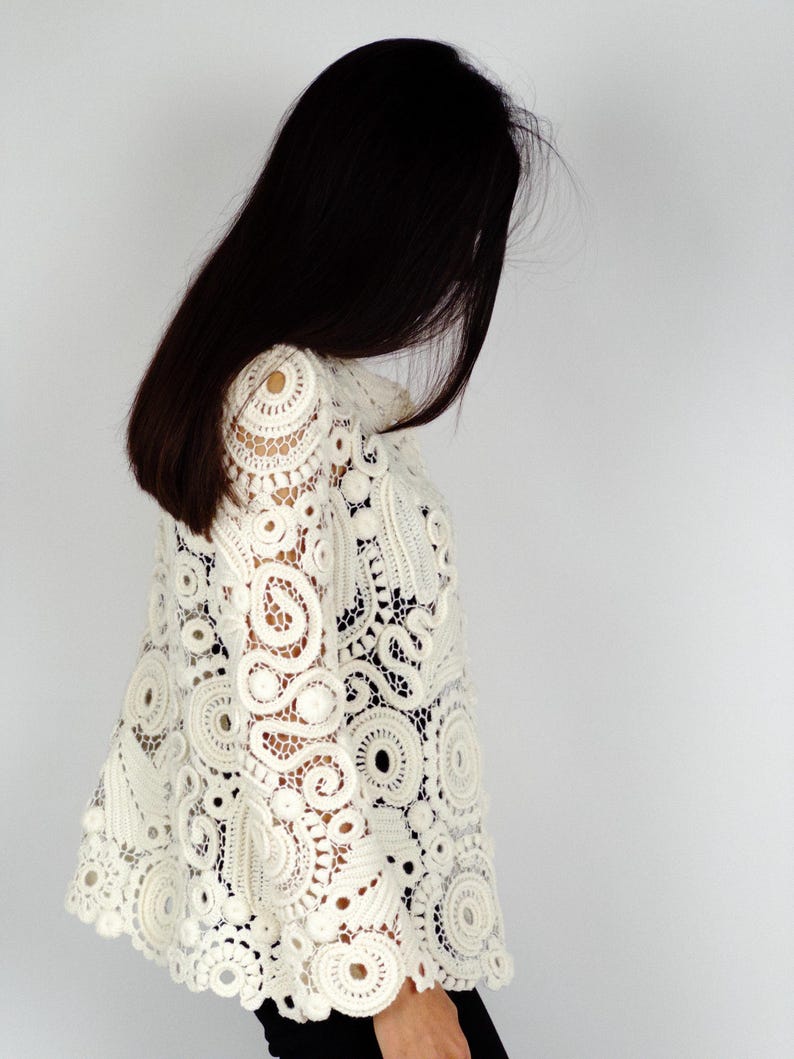 May include: Ivory-colored crochet top with intricate circular and floral patterns. The openwork design features a high collar and long sleeves. The top is made of a soft, lightweight material.