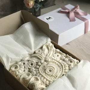 May include: A cream-colored, intricately crocheted item rests in an open cardboard box lined with tissue paper. A white gift box with a pink ribbon sits nearby. The text in the image reads: "Give yourself or a loved one something unforgettable."