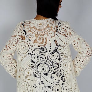 May include: Cream-colored, long-sleeved crochet jacket featuring intricate floral and circular patterns. The openwork design reveals the wearer's clothing underneath. The jacket has a round neckline and a slightly flared hem.