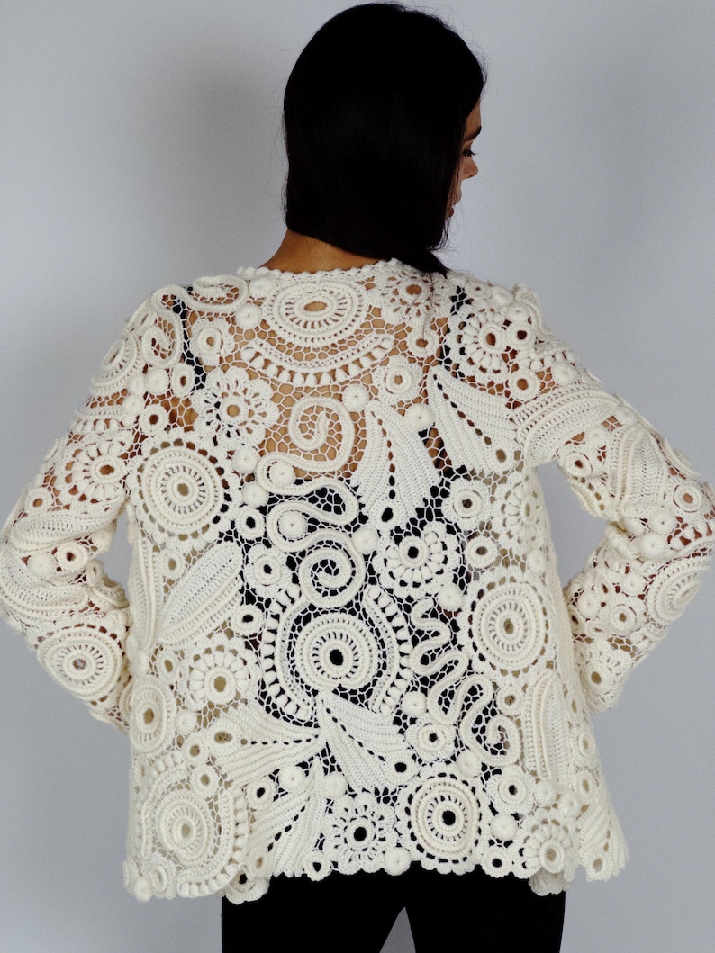 May include: Cream-colored, long-sleeved crochet jacket featuring intricate floral and circular patterns. The openwork design reveals the wearer's clothing underneath. The jacket has a round neckline and a slightly flared hem.