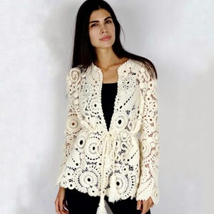 May include: Cream-colored, open-front crochet jacket with long sleeves. The jacket features a detailed, circular pattern and a drawstring waist with tassels. The garment is worn over a black top and paired with black pants.