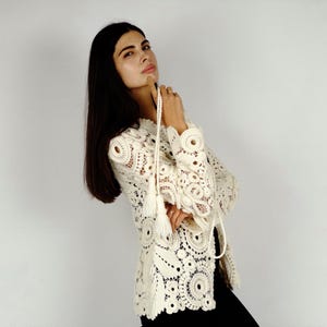 May include: Cream-colored crochet top with long sleeves and a decorative tassel tie. The top features intricate circular patterns and openwork details. The garment is worn with black pants. The style is bohemian and vintage-inspired.