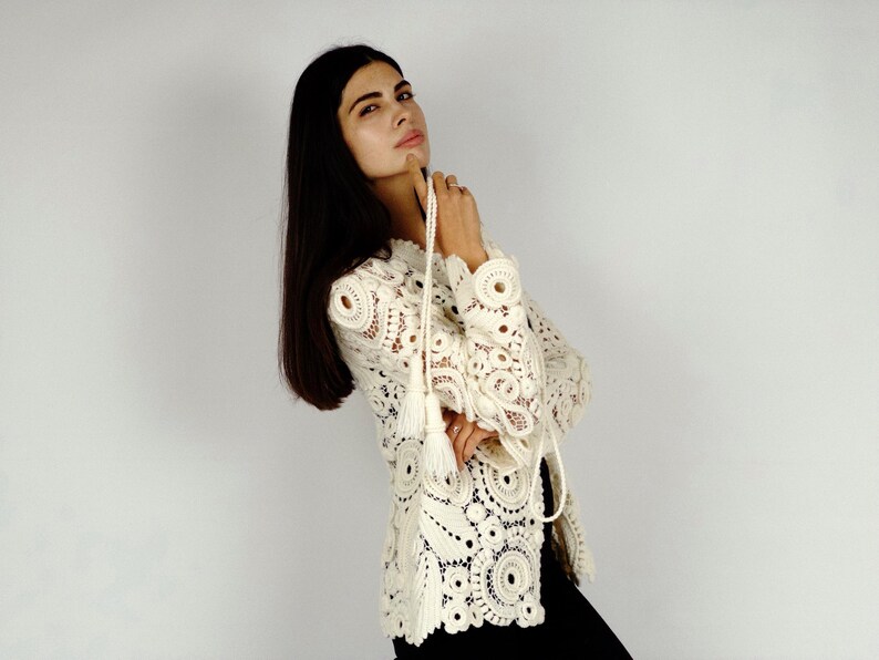 May include: Cream-colored crochet top with long sleeves and a decorative tassel tie. The top features intricate circular patterns and openwork details. The garment is worn with black pants. The style is bohemian and vintage-inspired.