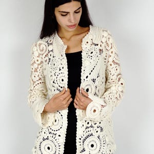 May include: Cream-colored, open-front crochet jacket with intricate circular and floral patterns. The jacket features long sleeves and a collarless design. The handmade garment is a delicate, lacy style, perfect for layering.