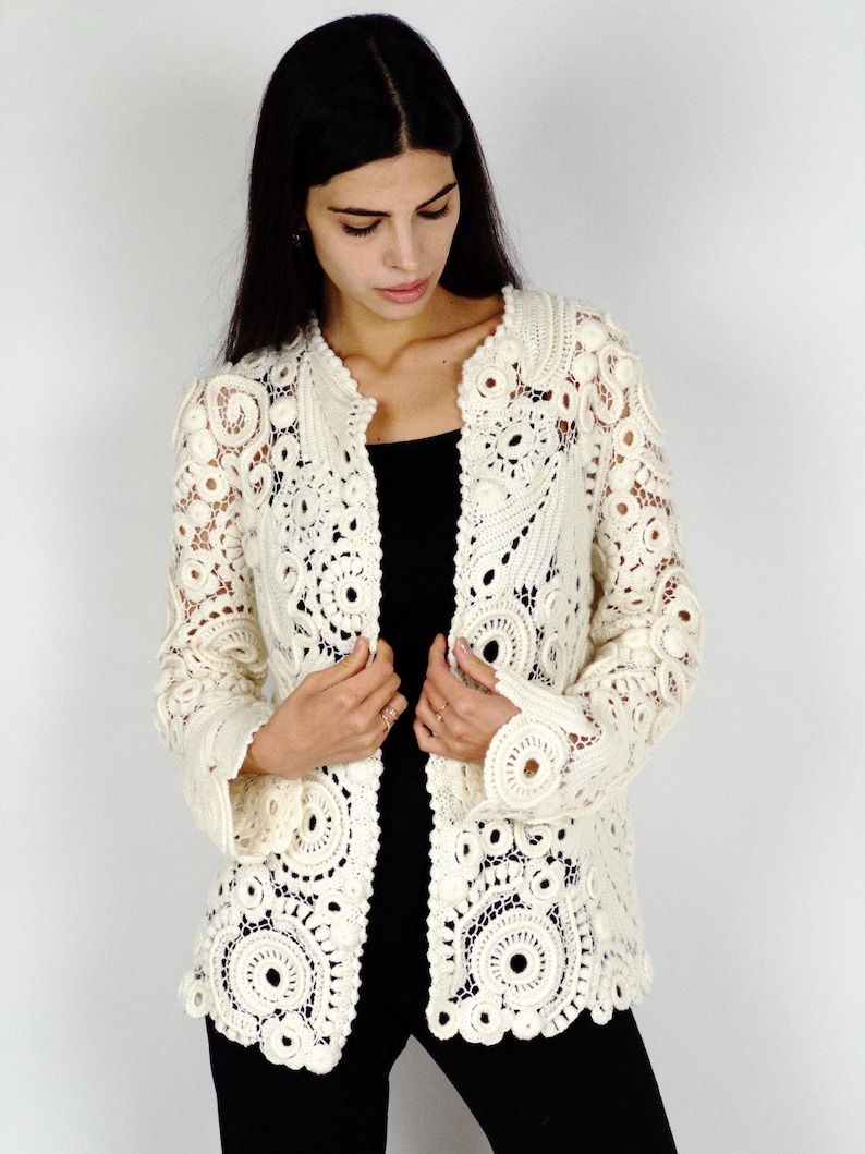 May include: Cream-colored, open-front crochet jacket with intricate circular and floral patterns. The jacket features long sleeves and a collarless design. The handmade garment is a delicate, lacy style, perfect for layering.