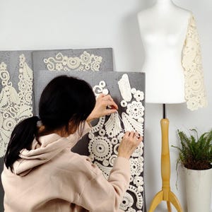 May include: A person working on a cream-colored lace sleeve and decorative panels. The panels are gray and feature intricate, circular lace designs. A dress form with a wooden stand and a potted plant are also visible.