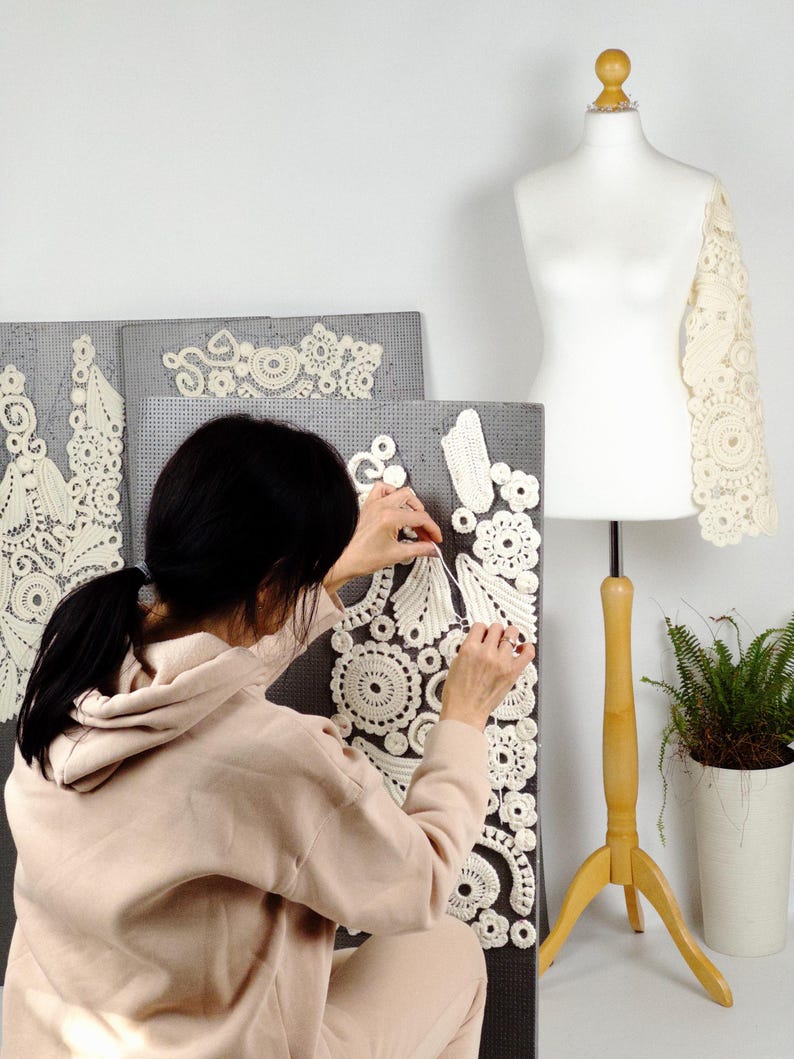 May include: A person working on a cream-colored lace sleeve and decorative panels. The panels are gray and feature intricate, circular lace designs. A dress form with a wooden stand and a potted plant are also visible.